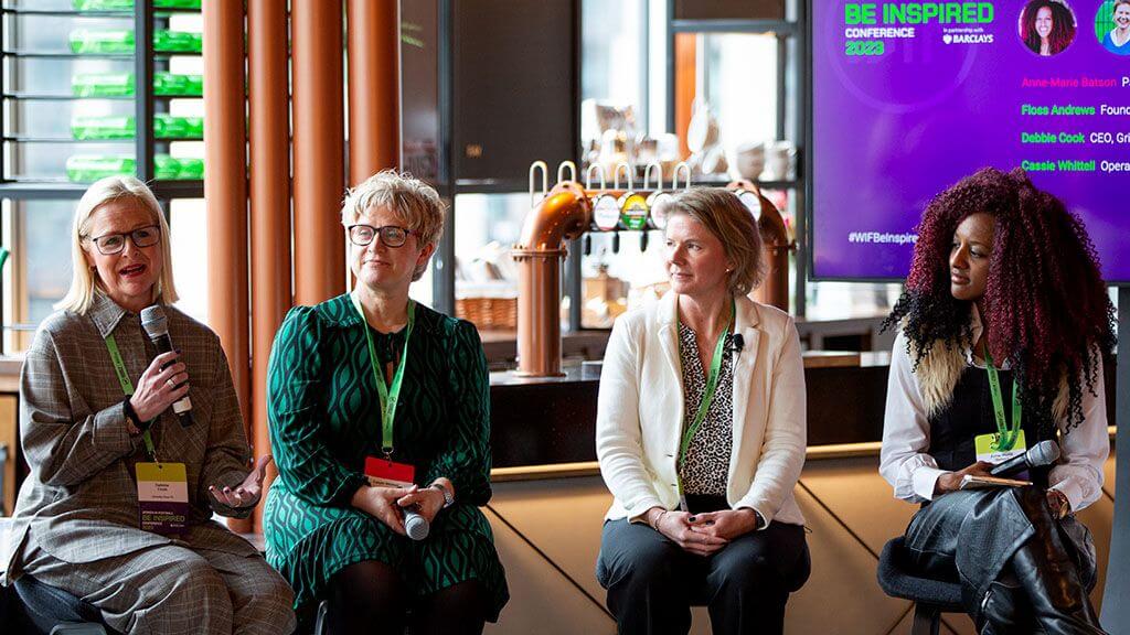 Four women on a panel at the Be Inspired Conference, seated on tall barstools, the leftmost speaking into a microphone